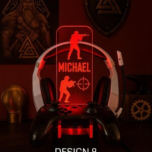 May include: A red LED gaming lamp with a personalized name and soldier silhouettes. The lamp sits on a black game controller base, with a white and black headset behind it. The text "DESIGN 8" is at the bottom.