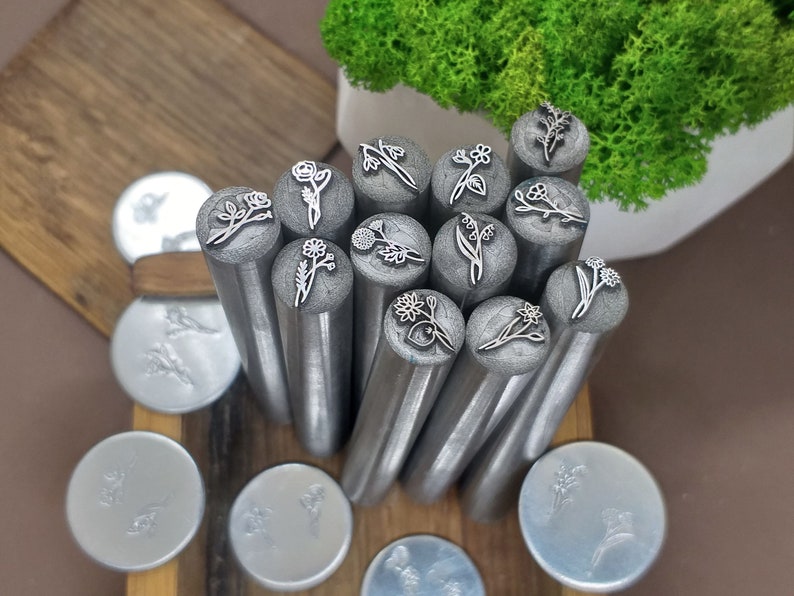 Puede incluir: Un conjunto de 15 cabezales de sello de flores de metal, dispuestos en c&iacute;rculo sobre una superficie de madera. Los cabezales de sello son plateados y tienen varios dise&ntilde;os florales.