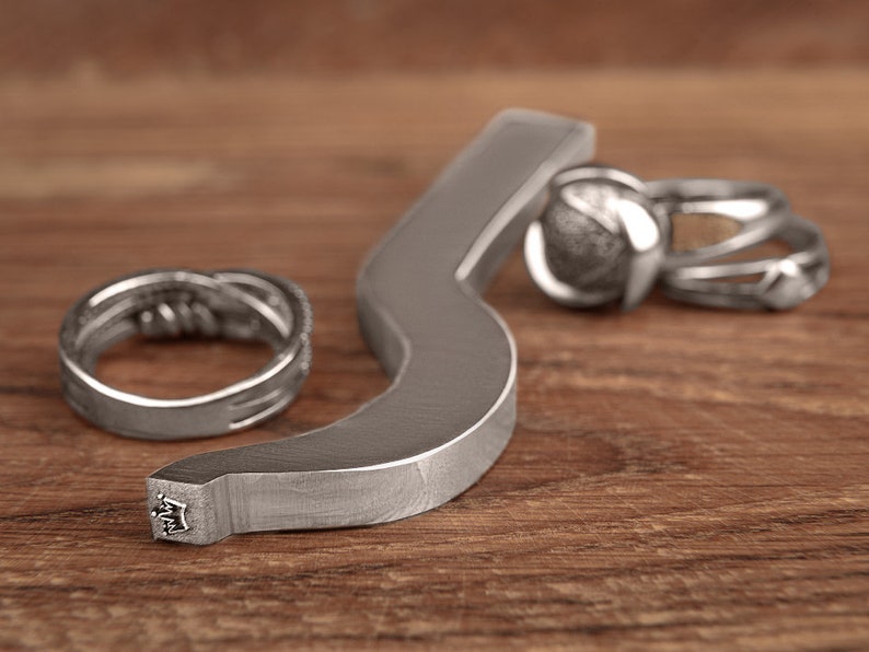 May include: A silver metal ring making tool with a crown design on the handle. The tool is lying on a wooden surface with three silver rings next to it.