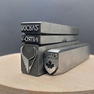 Pu&ograve; includere: Quattro timbri in metallo con diversi design. Il timbro superiore recita "80SCLAS", il secondo timbro recita "V-ORTII", il terzo timbro presenta una testa aliena stilizzata e il timbro inferiore presenta uno scudo con una foglia d'acero e il testo "CANADA".