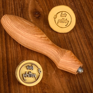 May include: A wooden handle with a metal screw attached to a round brass stamp with the text "love pottery" engraved on it. The stamp is on a wooden surface.