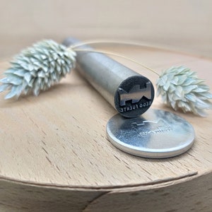 Pu&ograve; includere: Un timbro in metallo argentato con il testo "LEGO FUENTE" inciso sul lato. Il timbro &egrave; su una superficie di legno con fiori secchi.