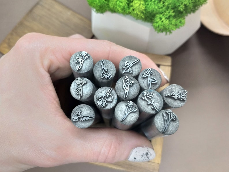 Puede incluir: Un conjunto de 12 sellos met&aacute;licos con diferentes dise&ntilde;os florales. Los sellos son de color plateado y tienen una forma redondeada. Est&aacute;n dispuestos en una mano, con los dise&ntilde;os hacia arriba.