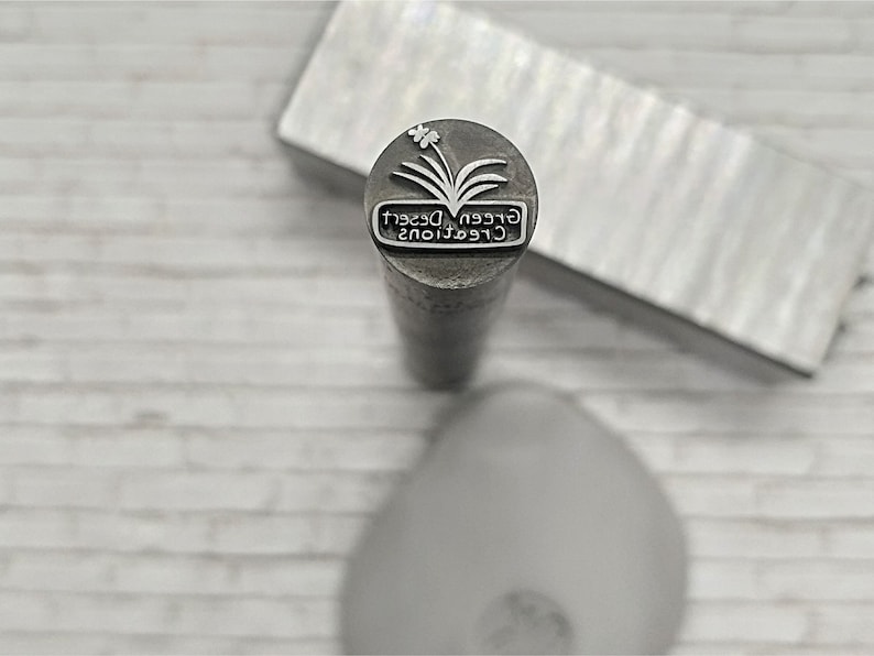 May include: A silver metal stamp with the text "Green Desert Creations" and a stylized image of a plant with a flower.