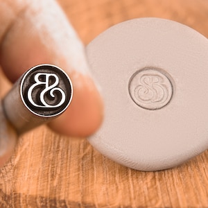 May include: A close-up of a metal stamp with the letters "&" engraved on the face. The stamp is being held by a hand and is being used to create an impression on a piece of clay.