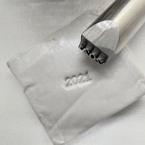 May include: A close-up of a silver metal stamp with the year 2021 engraved on it. The stamp is being used to imprint the year 2021 into a white clay surface.