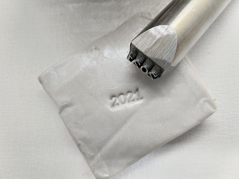 May include: A close-up of a silver metal stamp with the year 2021 engraved on it. The stamp is being used to imprint the year 2021 into a white clay surface.