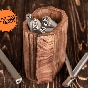 Pu&ograve; includere: Un set di timbri metallici con vari disegni, tra cui una bottiglia, un fiore e una lettera, sono esposti in un contenitore di legno. I timbri vengono utilizzati per creare disegni personalizzati su metallo.