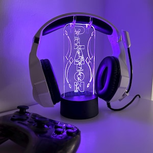 May include: White gaming headset with black earcups and a microphone, resting on a custom LED light stand. The stand displays the name "PLATON" in a glowing, stylized font. A black game controller is in the foreground.