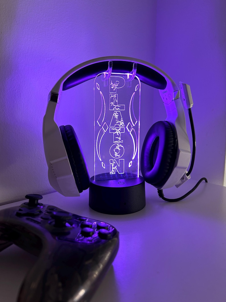 May include: White gaming headset with black earcups and a microphone, resting on a custom LED light stand. The stand displays the name "PLATON" in a glowing, stylized font. A black game controller is in the foreground.