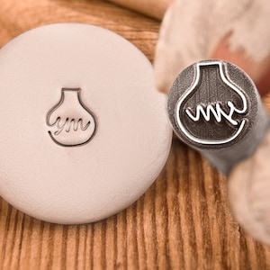 May include: A close-up of a metal stamp with a design of a lightbulb with the letters "ym" inside. The stamp is on a white surface.