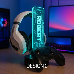 May include: A gaming setup featuring a white and black headset, a black game controller, and a personalized LED light with the name "ROBERT" in teal. The base of the light has the text "DESIGN 2". The background includes a computer monitor.