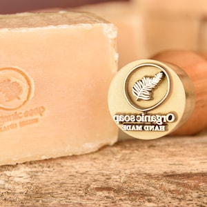 May include: A close-up of a wooden stamp with a gold metal top. The stamp has the words "HAND MADE ORGANIC SOAP" engraved on it. The stamp is next to a bar of soap with the words "ORGANIC SOAP HAND MADE" printed on it.