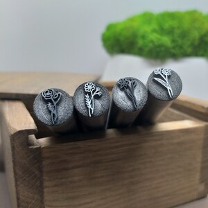 Puede incluir: Cuatro sellos de metal con dise&ntilde;os florales en una caja de madera. Los sellos son plateados y tienen diferentes dise&ntilde;os de flores, incluyendo una margarita, un narciso y una rosa.