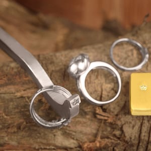 May include: A silver ring sizing tool with a silver ring and a gold ring with a crown design on a wooden surface.