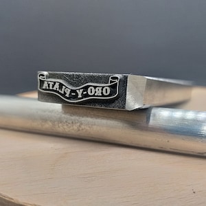 Pu&ograve; includere: Un timbro in metallo argentato con un design a banner che recita "ORO-Y-PLATA".