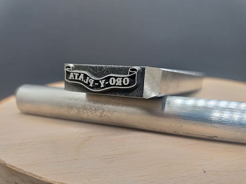 Pu&ograve; includere: Un timbro in metallo argentato con un design a banner che recita "ORO-Y-PLATA".