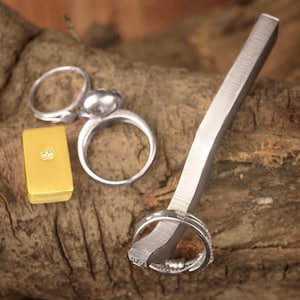 May include: Three silver rings on a wooden surface. One ring is a simple band, another is a band with a decorative design, and the third is a ring with a round, smooth top. A small gold box with a crown design is also on the surface.