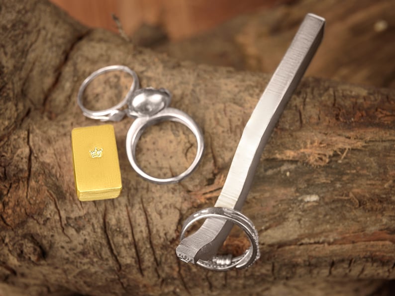 May include: Three silver rings on a wooden surface. One ring is a simple band, another is a band with a decorative design, and the third is a ring with a round, smooth top. A small gold box with a crown design is also on the surface.