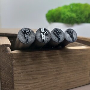Puede incluir: Cuatro sellos de flores de metal en una caja de madera. Los sellos tienen la forma de diferentes flores, incluyendo una rosa, una margarita, una flor silvestre y un narciso.