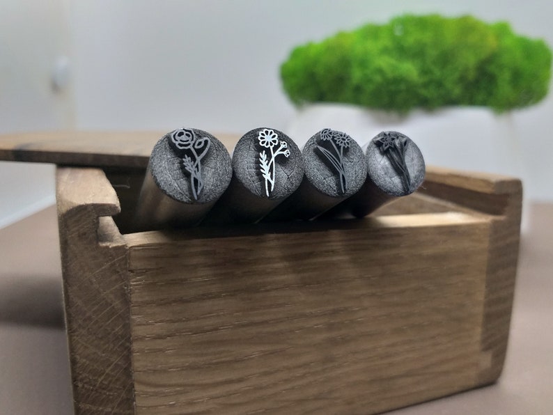 Puede incluir: Cuatro sellos de flores de metal en una caja de madera. Los sellos tienen la forma de diferentes flores, incluyendo una rosa, una margarita, una flor silvestre y un narciso.