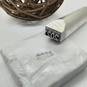 May include: A silver metal stamp with the year 2021 engraved on it. The stamp is used to create an impression on a white surface, which is also shown in the image.