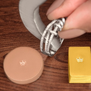 May include: A close-up of a silver ring with a diamond-like design being measured with a ring sizer. A small gold bar with a crown design is in the background.