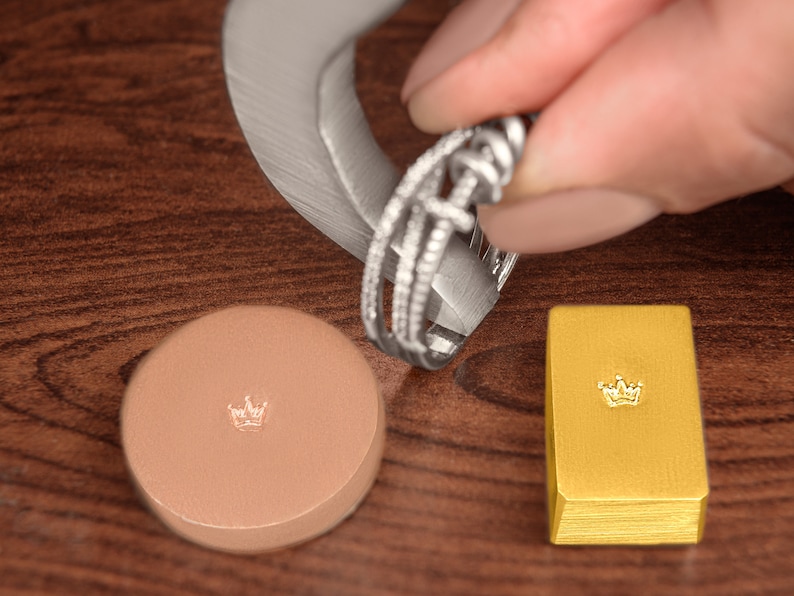 May include: A close-up of a silver ring with a diamond-like design being measured with a ring sizer. A small gold bar with a crown design is in the background.