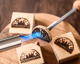May include: A wood burning tool with a wooden handle and a metal shaft is being used to brand a wooden block with the words "WOODWORKS" and a design of trees. The tool is being heated with a torch.