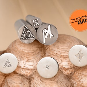 May include: Five metal stamps with different designs. The stamps are arranged on a wooden surface. The designs include a triangle with a circle inside, the letters "HK", the letters "PA", and a stylized leaf. The stamps are labeled "Custom Made".