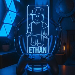 May include: A blue-lit gaming accessory featuring a cartoon character with the name "ETHAN" and the text "DESIGN 4". The design is set on a game controller base, with headphones and geometric wall panels in the background.