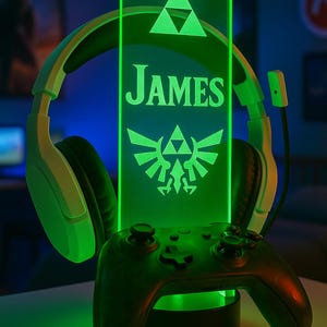 Peut inclure: Lampe LED verte avec le nom "JAMES" et une manette de jeu. La lampe présente un design avec des triangles et un emblème stylisé. Un casque blanc et vert est en arrière-plan. Le texte "DESIGN 4" est en bas.