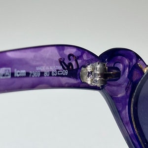 May include: Purple plastic sunglasses with a silver hinge and blue lenses. The sunglasses have the text "Made in Austria PA Icm 7369 80 63 009" printed on the frame.