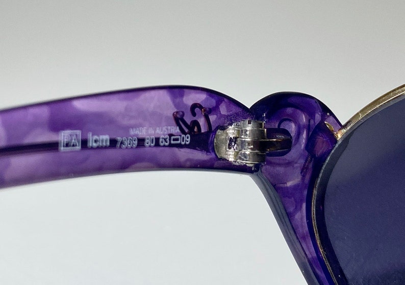 May include: Purple plastic sunglasses with a silver hinge and blue lenses. The sunglasses have the text "Made in Austria PA Icm 7369 80 63 009" printed on the frame.