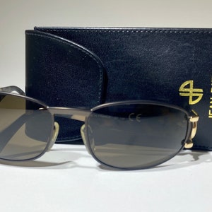 May include: Oval-shaped sunglasses with a dark frame and gradient lenses, next to a black leather case. The case has gold lettering that reads "JEAN PATOU PARIS" and a gold logo.