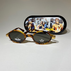 May include: Tortoise shell oval sunglasses with a gold trim and Blue Bay logo on the lenses. The sunglasses are UV protected 400 NM. The sunglasses are in a black case with a band of musicians on it.
