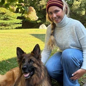 May include: A woman wearing a burgundy and gold striped knit headband, a gray turtleneck sweater, and blue jeans. A long-haired German Shepherd dog is lying on the grass next to her. The dog has brown and black fur.