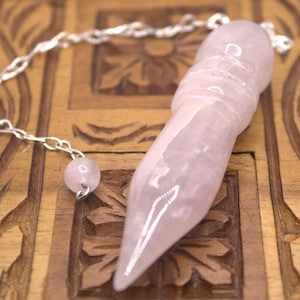 May include: A pink rose quartz pendulum with a silver chain and a small pink bead. The pendulum is on a wooden surface.