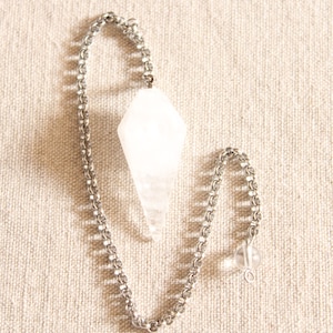 Peut inclure: Un pendule en cristal blanc avec une chaîne argentée. Le pendule a la forme d'une larme et se termine en pointe. La chaîne est composée de petits anneaux métalliques liés.