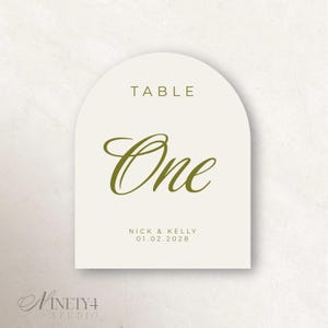 May include: An arched table number card in off-white with olive green text. The card features the word TABLE in a sans-serif font, the word One in a large script font, and the names NICK & KELLY with the date 01.02.2028 printed at the bottom.