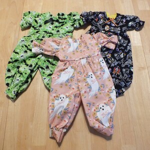 May include: Three Halloween-themed baby jumpsuits. One is green with bats and ghosts, one is black with skeletons, and one is pink with white ghosts and flowers.
