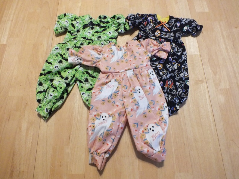 May include: Three Halloween-themed baby jumpsuits. One is green with bats and ghosts, one is black with skeletons, and one is pink with white ghosts and flowers.
