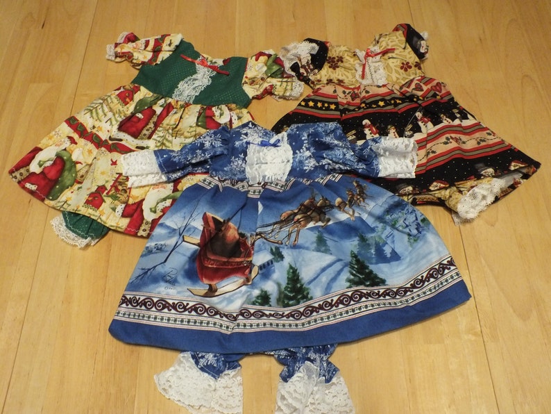 May include: Three Christmas-themed doll dresses. One dress is green and white with a red and white Santa Claus print. Another dress is blue and white with a winter scene featuring a sleigh and reindeer. The third dress is black, red, and white with a snowman and Santa Claus print.