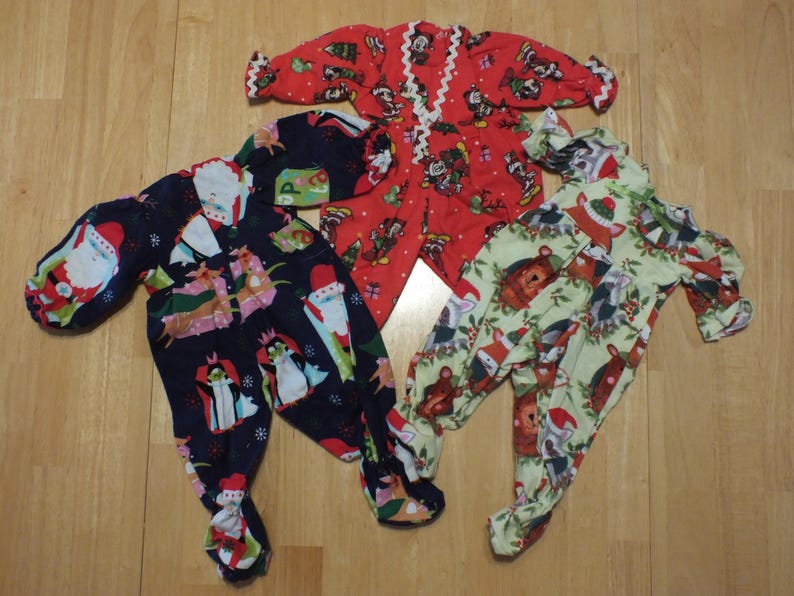 May include: Three pairs of Christmas pajamas for babies. One pair is red with a white trim and features a Mickey Mouse design. Another pair is green with a white trim and features a woodland animal design. The last pair is blue with a white trim and features a Santa Claus design.