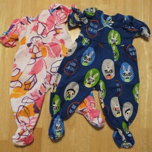 May include: Two pairs of baby pajamas. One pair is pink with orange and purple floral print. The other pair is blue with a white bunny print and the text "Too cool to be cute".
