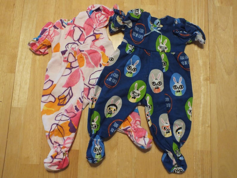 May include: Two pairs of baby pajamas. One pair is pink with orange and purple floral print. The other pair is blue with a white bunny print and the text "Too cool to be cute".