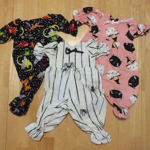 May include: Three baby Halloween pajamas. One is black with a space theme and dinosaurs, one is pink with black cats and witches hats, and one is white with black stripes and spiders.