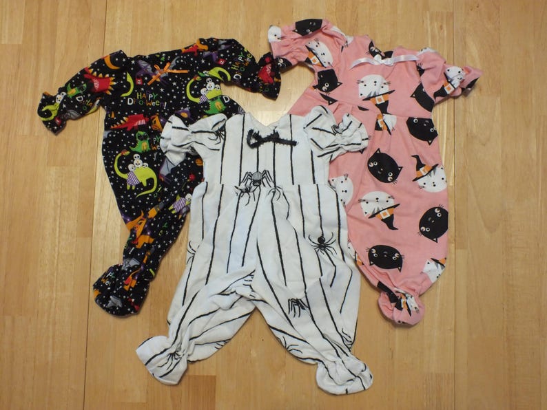 May include: Three baby Halloween pajamas. One is black with a space theme and dinosaurs, one is pink with black cats and witches hats, and one is white with black stripes and spiders.