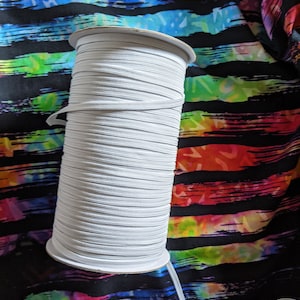 Pode incluir: Um carretel branco de cordão elástico, enrolado firmemente em um núcleo de papelão. O elástico está sobre um tecido colorido com listras pretas.