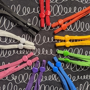 Puede incluir: Una colección de ajustadores de cordón elástico de colores, en negro, blanco, rojo, naranja, amarillo, verde, rosa, morado y azul. Cada ajustador tiene un botón para ajustar la longitud del cordón. El fondo es una tela negra con espirales blancas.